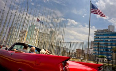 Turistas, que van en un convertible, pasan junto a la bandera de Estados Unidos y una bandera cubana colocadas en la Embajada estadounidense en La Habana. AFP
